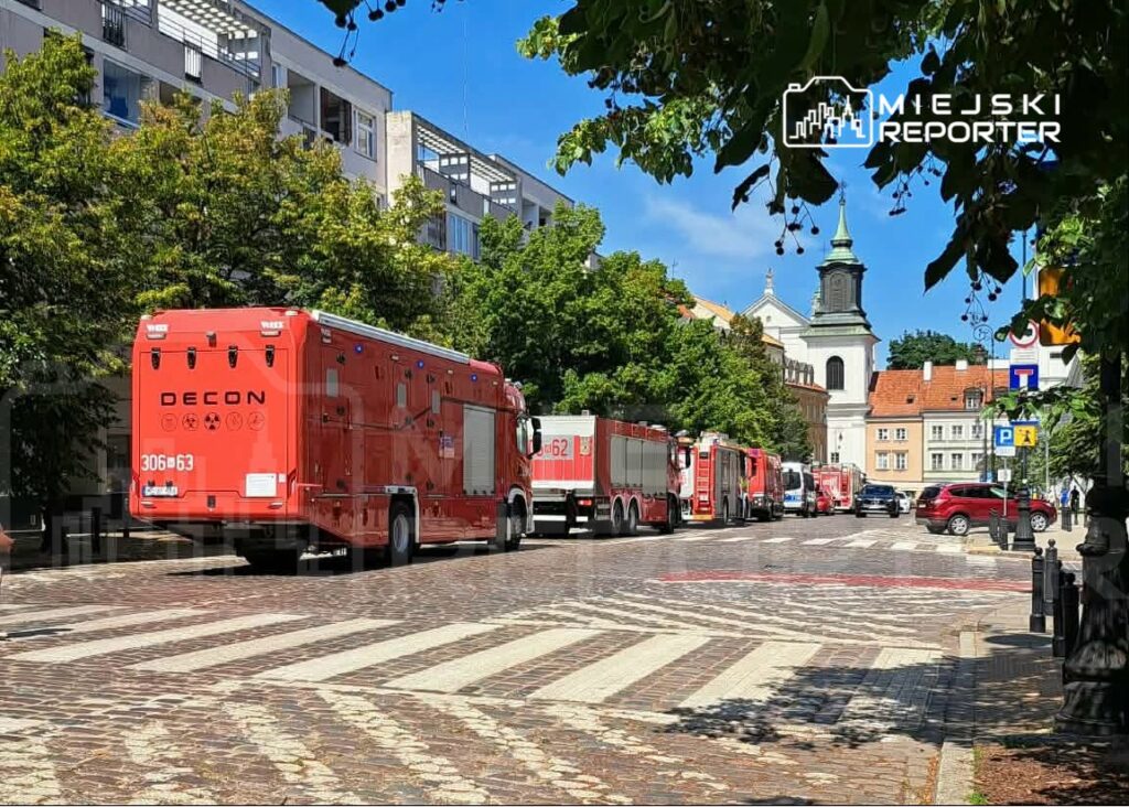 Czerwone wozy strażackie zaparkowane wzdłuż brukowanej ulicy, w tle widoczny kościół z wieżą.