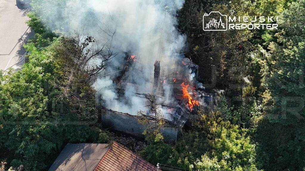 Płonąca konstrukcja otoczona dymem i roślinnością, w tle widoczna ulica oraz sąsiedni budynek.