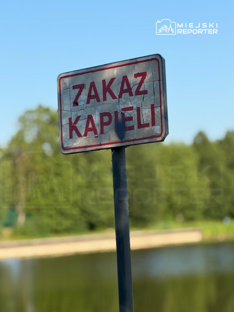 Zardzewiały znak "ZAKAZ KAPIELI" umieszczony nad brzegiem wody w otoczeniu zieleni.