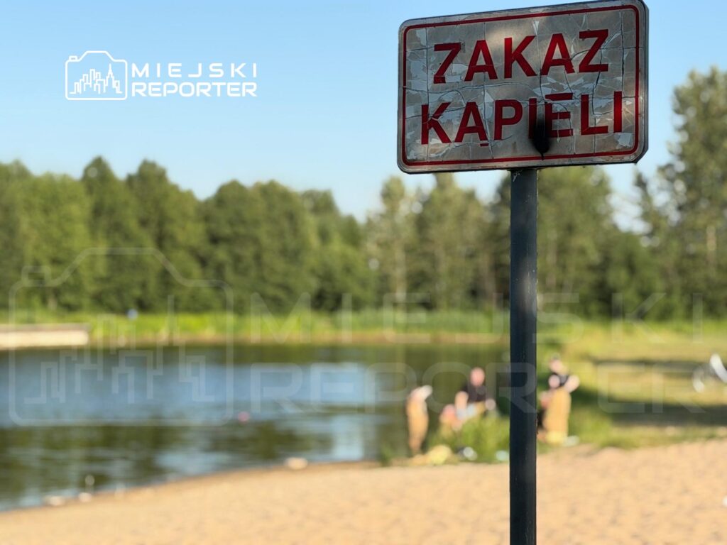 Znakiem „ZAKAZ KAPIELI” oznaczone jest miejsce nad wodą, w tle widoczni ludzie na plaży.