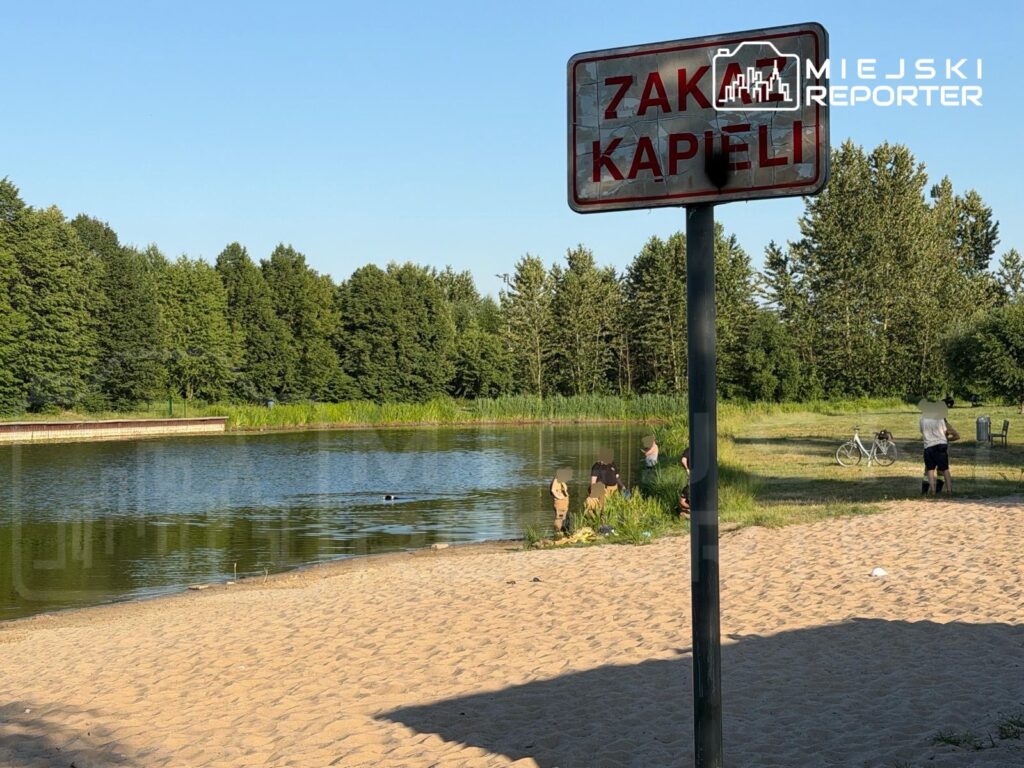Znakiem "ZAKAZ KAPIELI" oznaczony brzeg stawu, przy którym kilka osób stoi na piasku i w wodzie. W tle widać zieleń drzew.