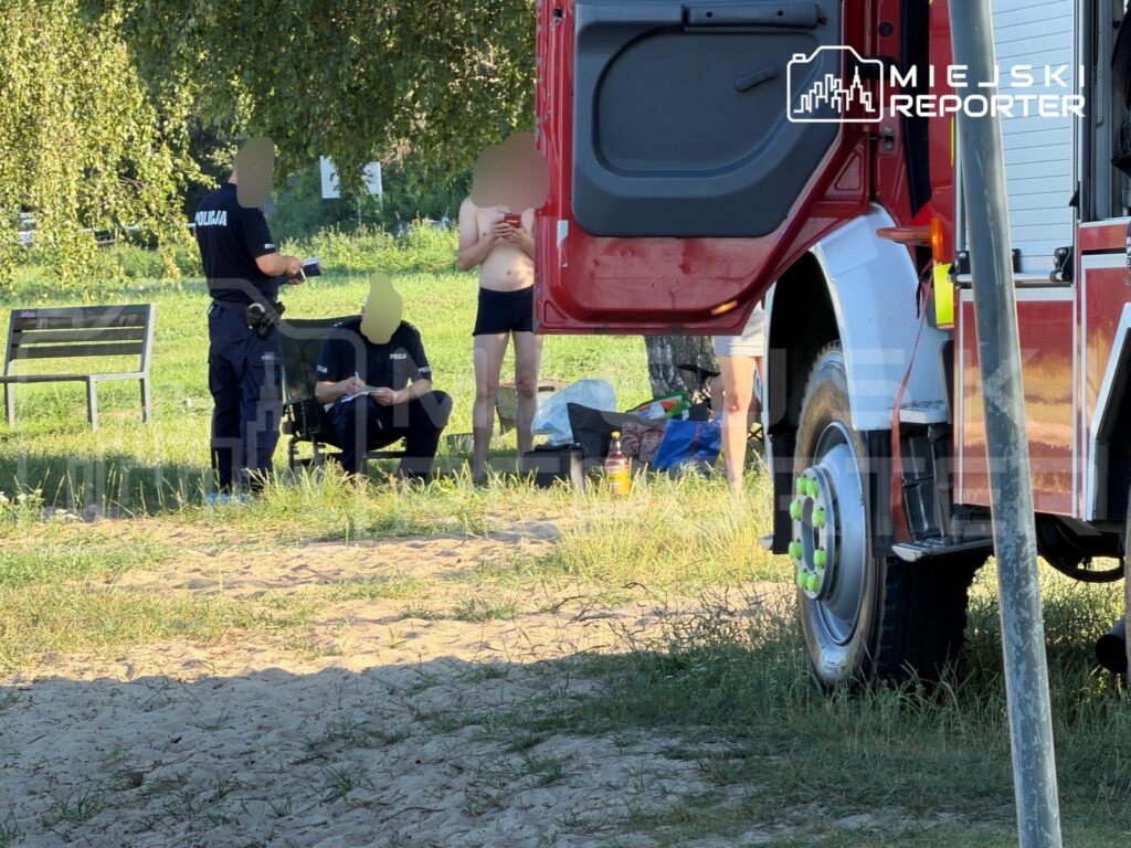 Mężczyzna w kąpielówkach rozmawia z policjantem, obok siedzi drugi funkcjonariusz, w tle widać samochód strażacki.