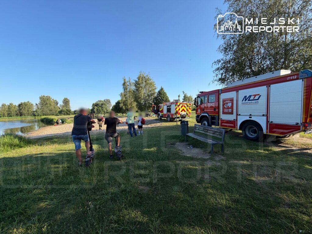 Grupa osób na hulajnogach zmierza w stronę wody, obok parkują wozy strażackie w słonecznym otoczeniu.
