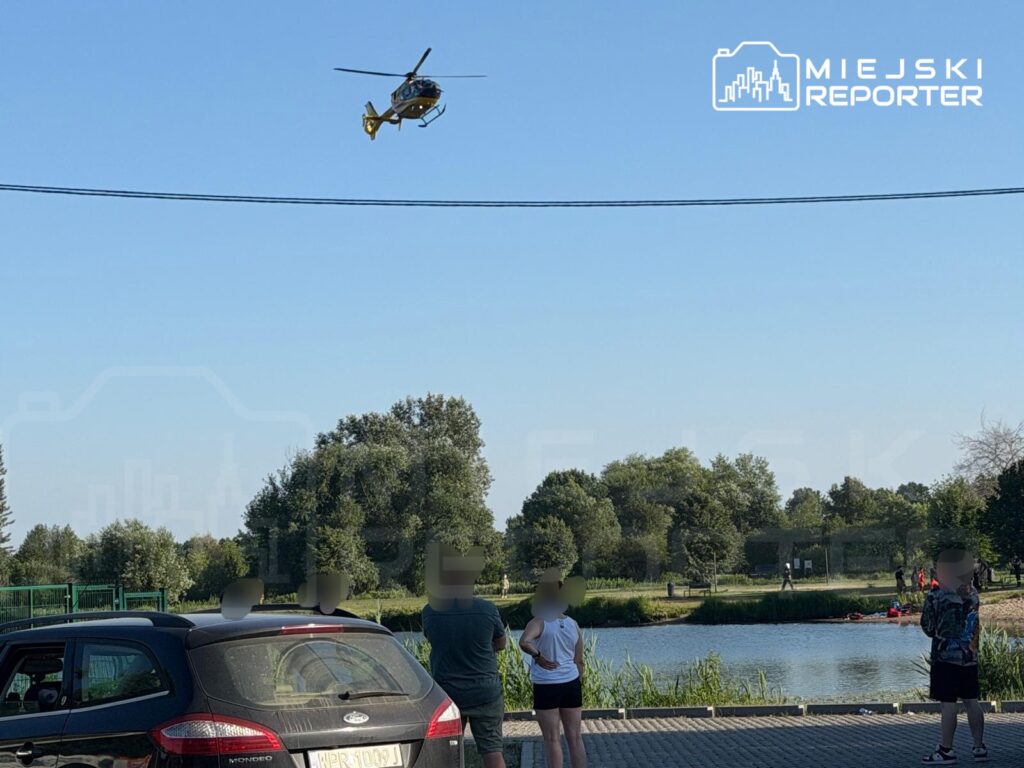 Helikopter przelatuje nad jeziorem, a grupa ludzi obserwuje go z brzegu, obok zaparkowanego samochodu.