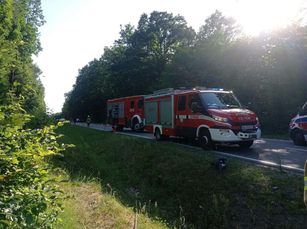 Wzdłuż drogi stoją dwa wozy strażackie, a strażacy pracują nad usunięciem przeszkody w lesie.