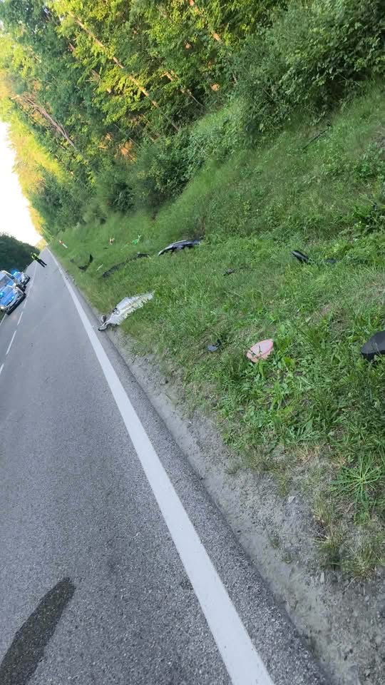 Na poboczu drogi leżą fragmenty pojazdu, obok widoczne są zarośla i drzewa. W tle stoi samochód policyjny.