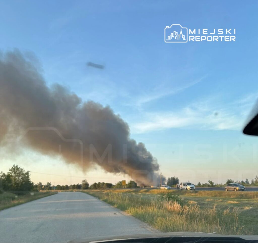 Gęsty czarny dym unoszący się z pożaru w oddali, widoczny z asfaltowej drogi otoczonej zielonymi polami.