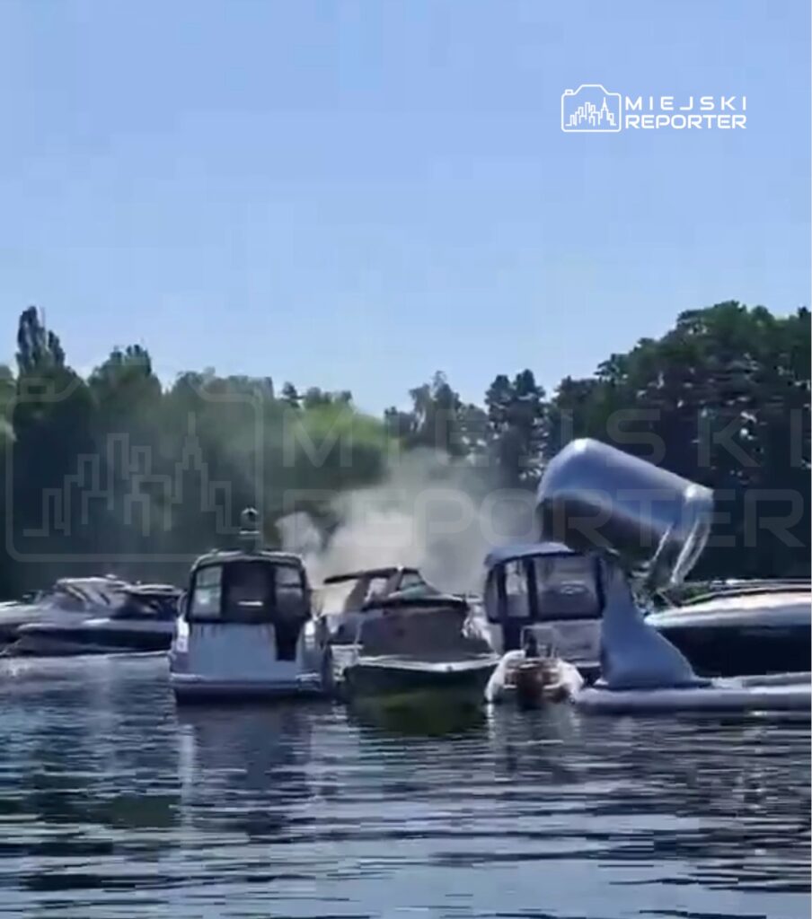Na wodzie przycumowane motorówki, z jednej z nich unosi się dym, a w tle zielone drzewa.