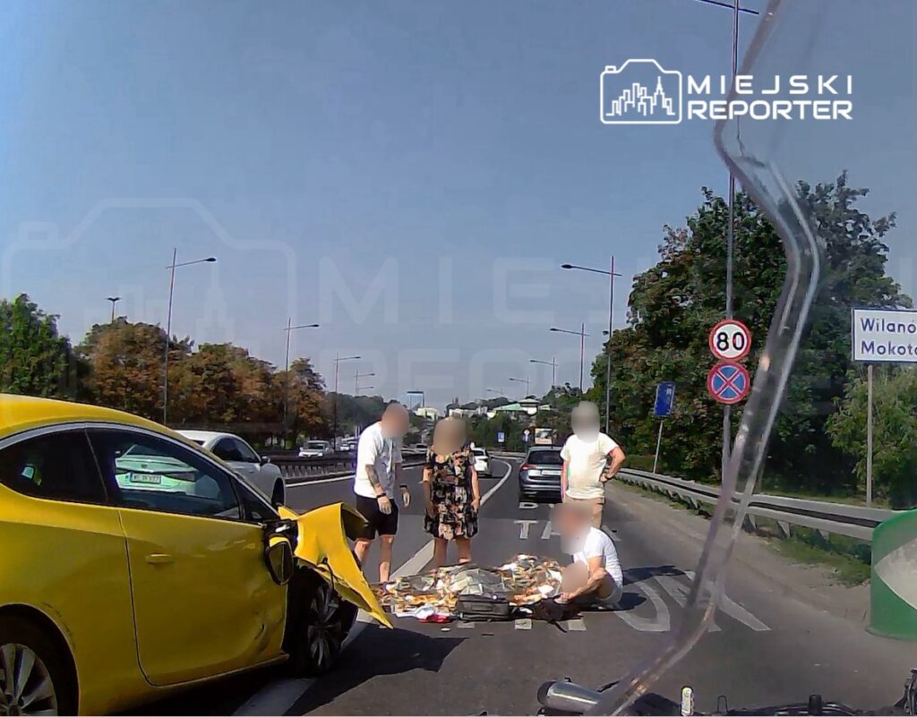 Żółty samochód osobowy z uszkodzoną stroną stoi na jezdni, a obok grupka ludzi zbiera rozsypane przedmioty.
