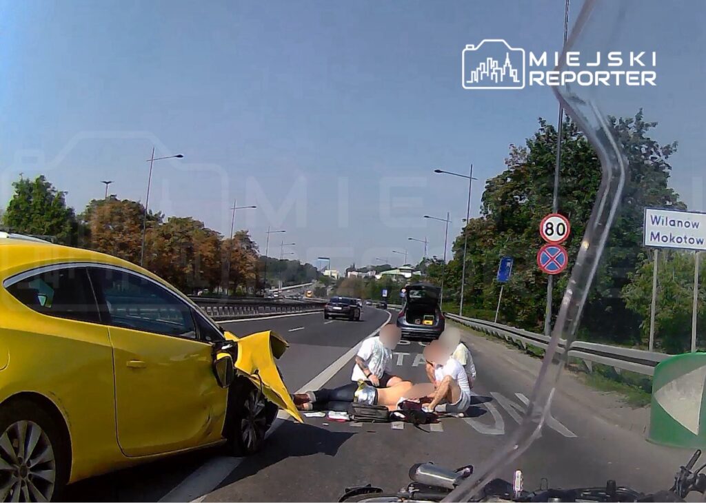 Na drodze, obok uszkodzonego żółtego samochodu, dwie osoby udzielają pomocy poszkodowanemu.