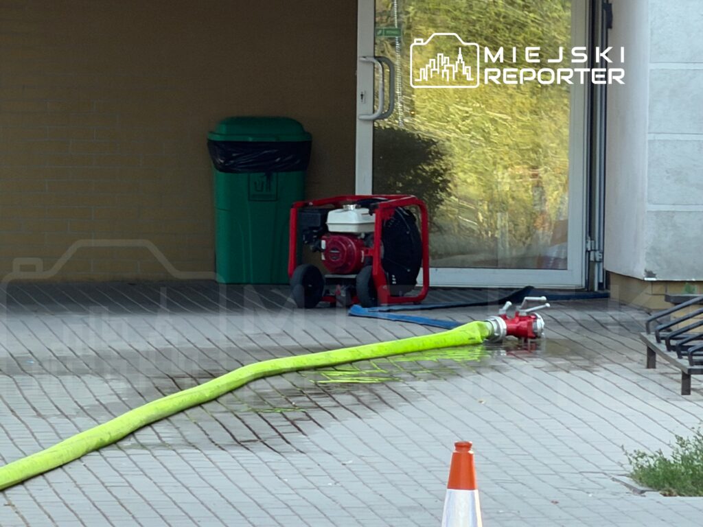Czerwony agregat prądotwórczy obok żółtej węża strażackiego na mokrej posadzce przed wejściem do budynku.