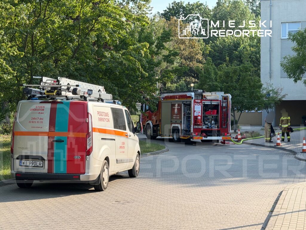 Samochód strażacki oraz pojazd pogotowia energetycznego zaparkowane w pobliżu budynku, z pracownikami przygotowującymi sprzęt.
