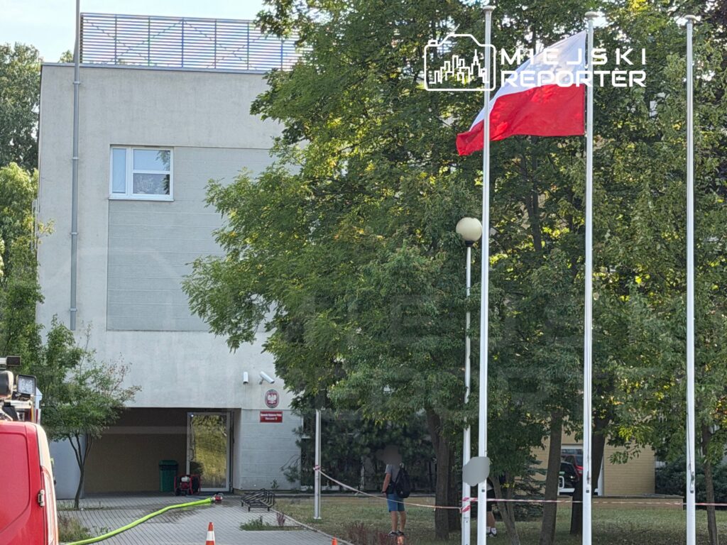Mężczyzna stoi obok flagi Polski przed nowoczesnym budynkiem, w tle widoczne drzewa i sprzęt budowlany.
