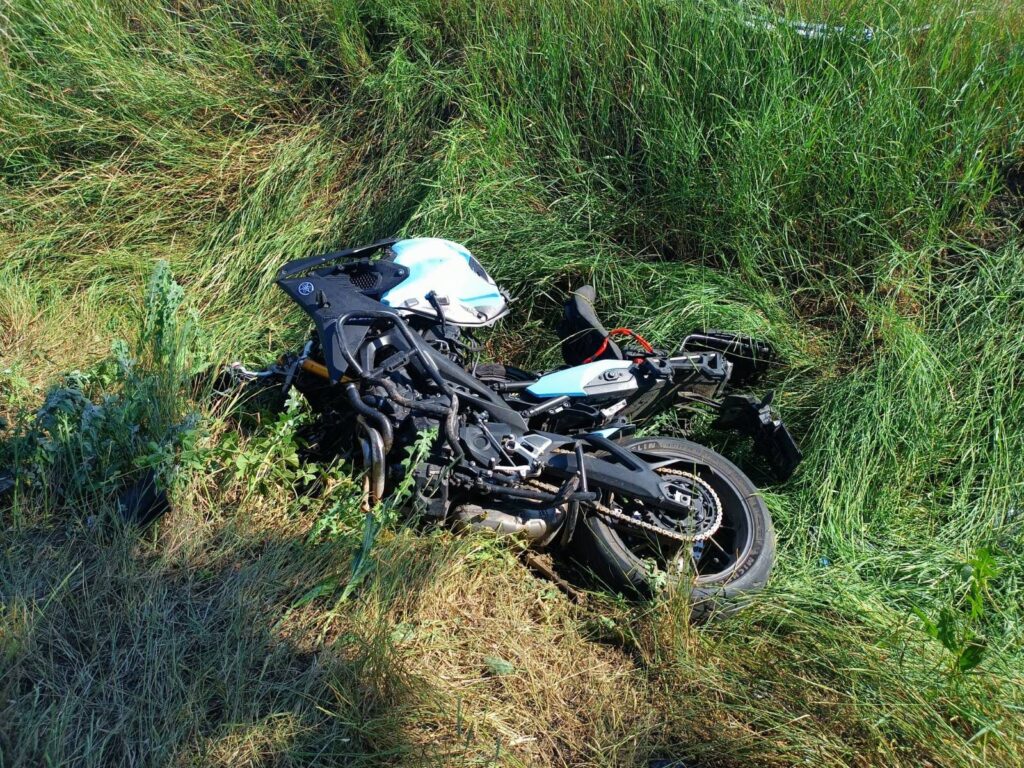 Złamany motocykl leży w wysokich trawach na poboczu drogi.