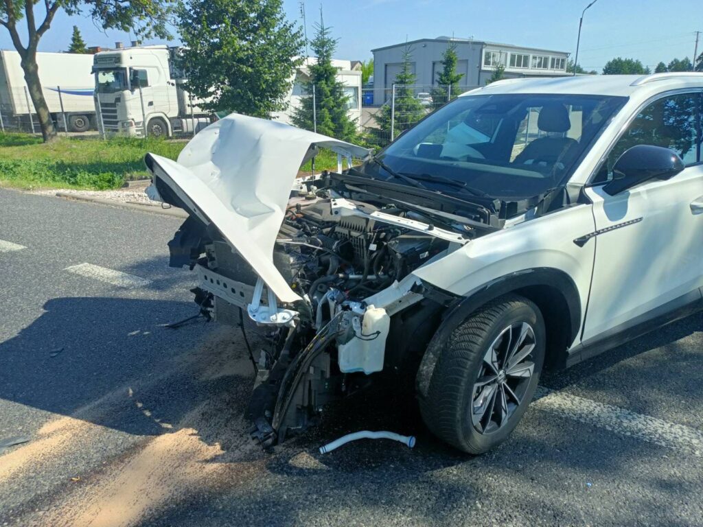 Uszkodzony samochód osobowy z otwartą maską stoi na drodze, w tle widoczny ciężarówka i zieleń.