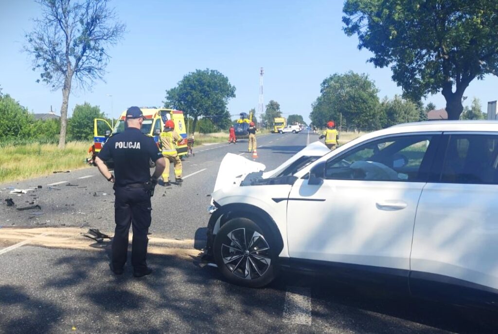 Policjant w mundurze obserwuje miejsce wypadku drogowego, gdzie zderzyły się dwa pojazdy. W tle widać pojazdy ratunkowe.