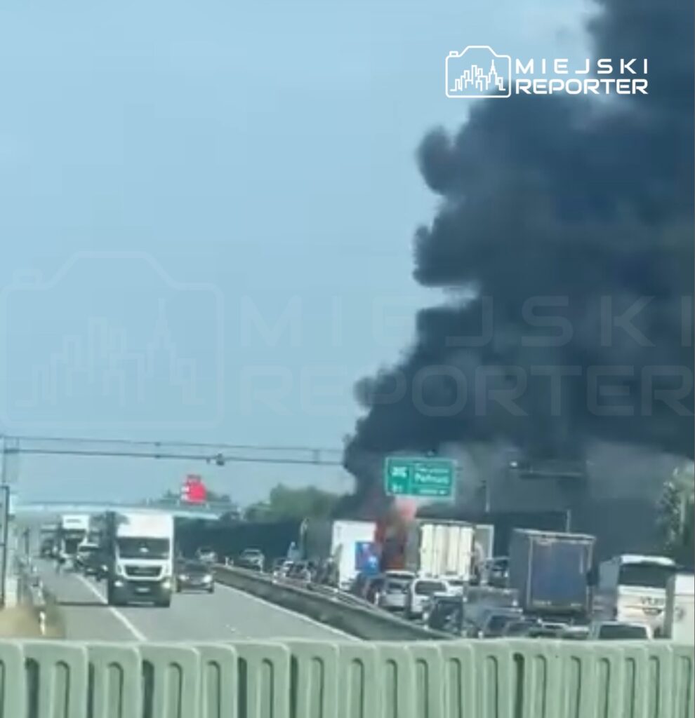 Gęsty czarny dym unoszący się nad zablokowaną autostradą, w tle widoczne ciężarówki i samochody osobowe.