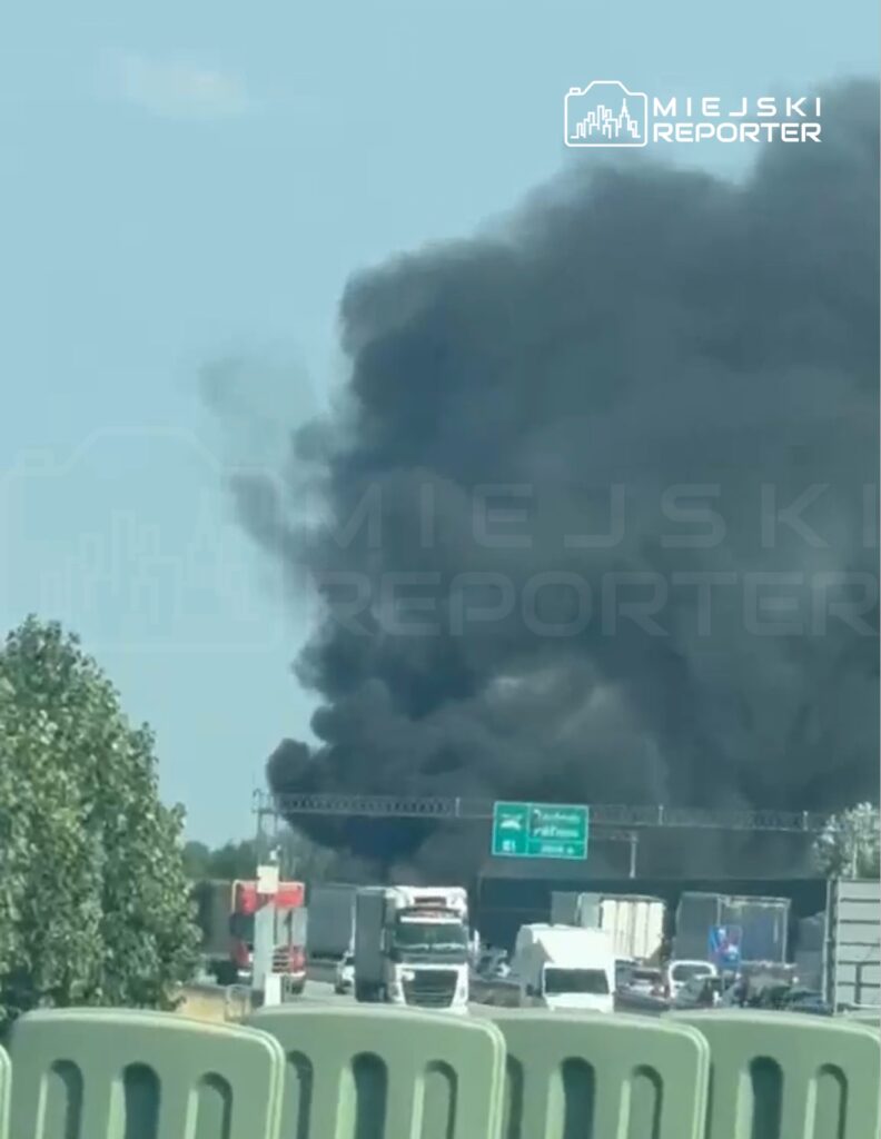 Gęsty czarny dym unoszący się z miejsca pożaru w pobliżu ruchliwej drogi, obok stojących ciężarówek.