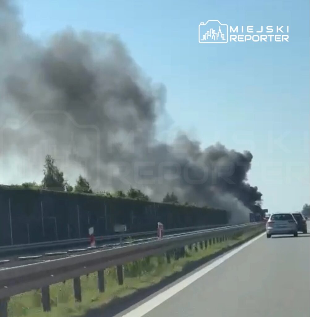 Gęsty czarny dym unosi się nad drogą, obok której znajdują się samochody i zieleń w tle.