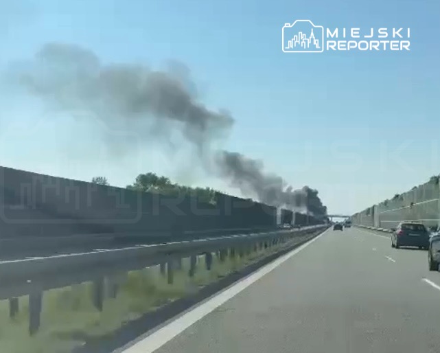 Na autostradzie widać unoszący się czarny dym z kierunku lasu, obok przejeżdżające samochody.