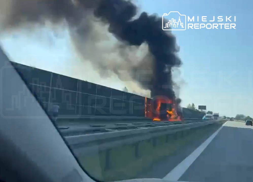 Na drodze, w pobliżu bariery, płonący pojazd wydobywa gęsty czarny dym.
