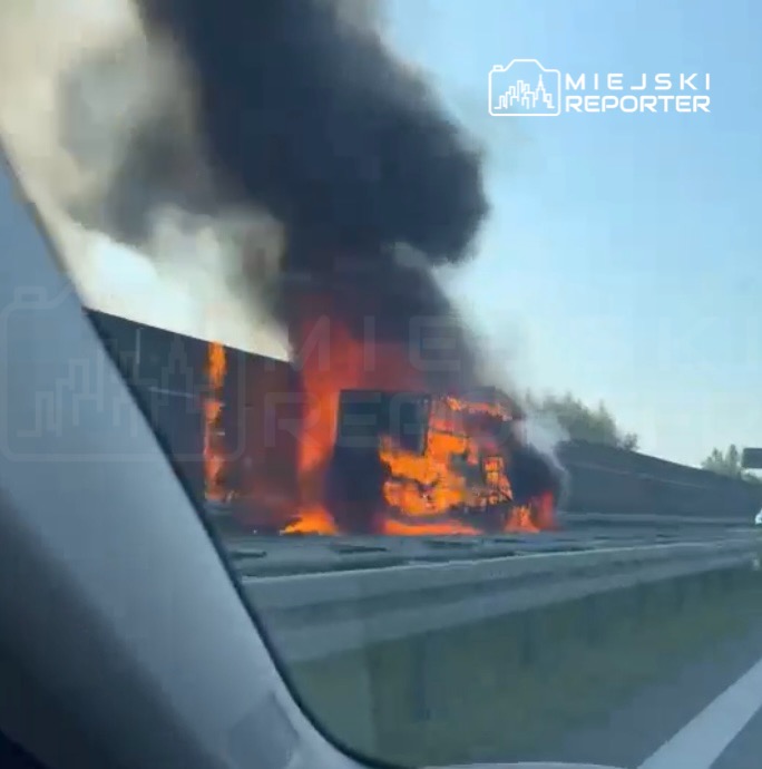 Pojazd płonący na autostradzie, w tle widoczne kłęby czarnego dymu.