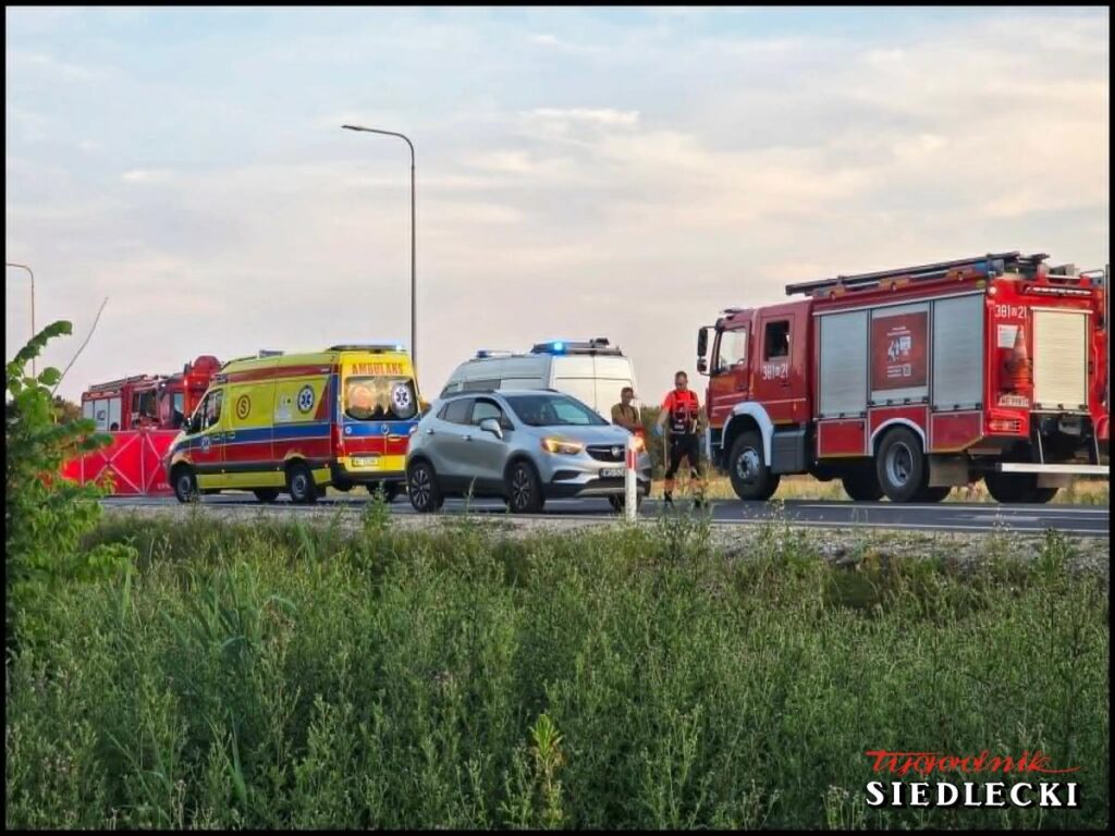 Na drodze stoją pojazdy służb ratunkowych, w tym karetka i wóz strażacki, obok dwóch ratowników rozmawiających z kierowcą.