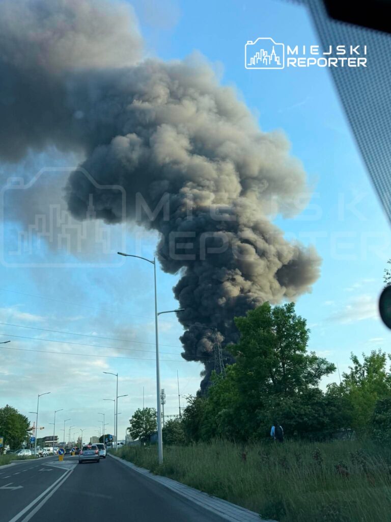Gęsty, czarny dym unoszący się w niebo z kierunku przemysłowego, widoczny z ruchliwej drogi.