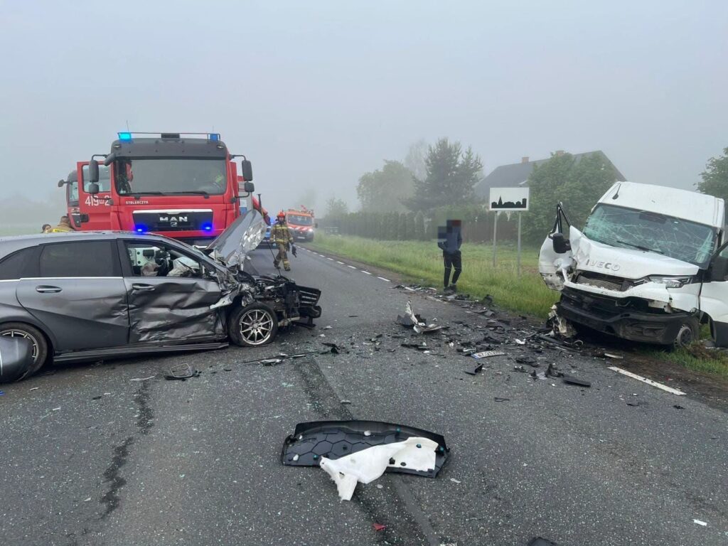 Zderzenie dwóch samochodów na drodze, z uszkodzonymi pojazdami i rozrzuconymi fragmentami karoserii. W tle widoczny wóz strażacki.