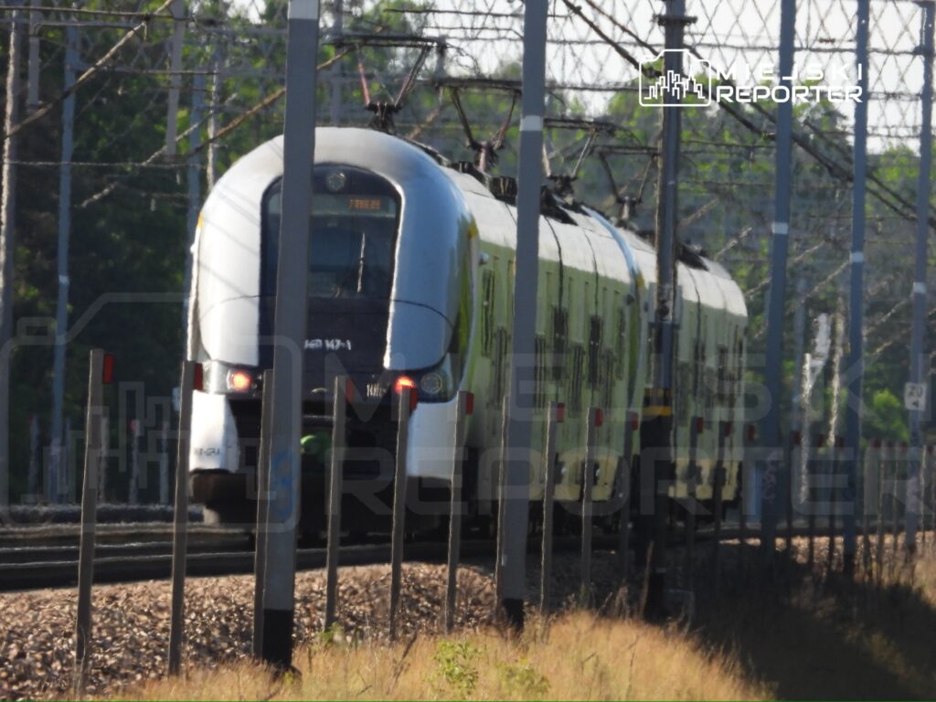 Pociąg elektryczny porusza się po torach w otoczeniu zieleni i słupów trakcyjnych.