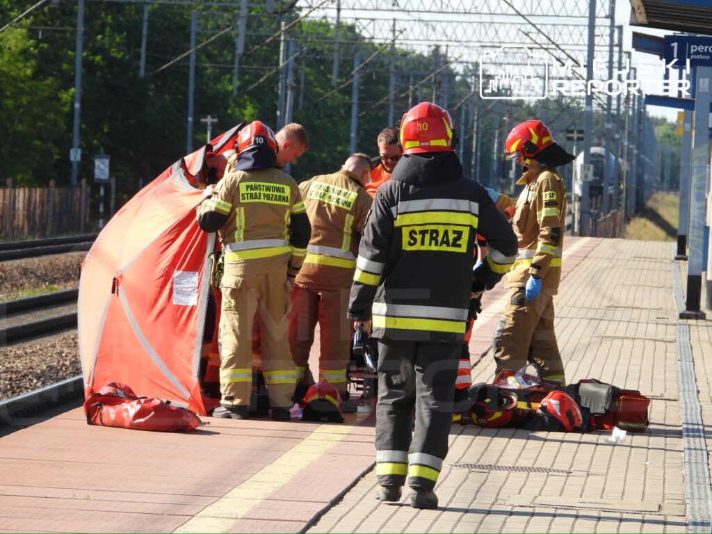 Grupa strażaków w mundurach pracuje nad osobą poszkodowaną na stacji kolejowej, otoczona pomarańczowym namiotem.