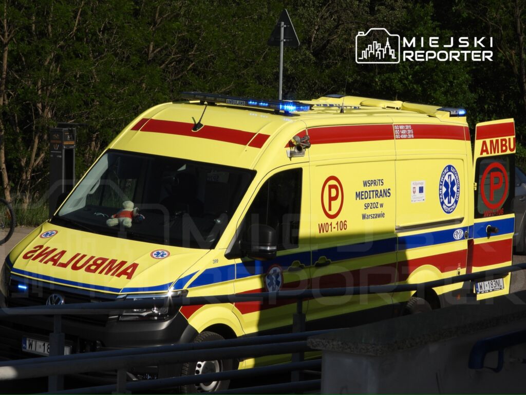 Żółta karetka pogotowia z niebieskimi światłami zaparkowana przy drodze, z pluszowym misiem na desce rozdzielczej.