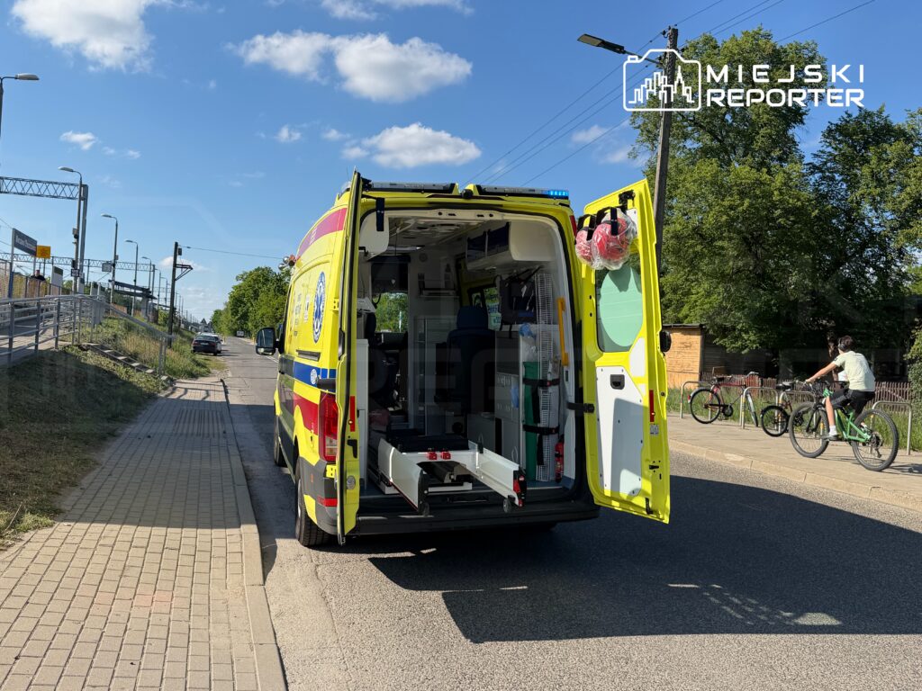 Ambulans z otwartymi drzwiami stoi przy ulicy, obok rowerzysty przejeżdżającego obok zaparkowanych rowerów.