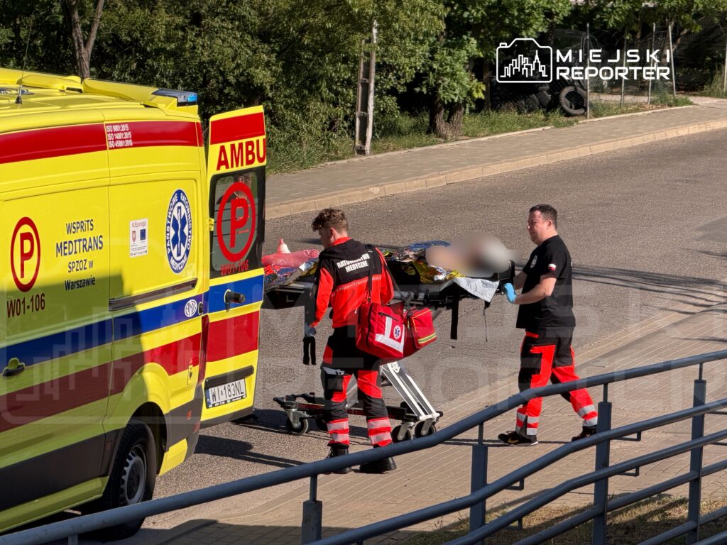 Ratownicy medyczni przenoszą nosze z pacjentem z karetki na ulicy w otoczeniu zieleni.