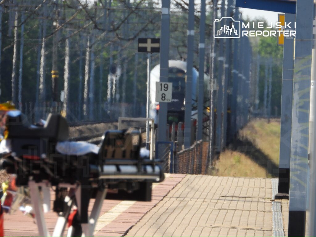 Pojazd szynowy zbliża się do peronu, na którym stoi wózek medyczny. W tle widoczne znaki kolejowe.