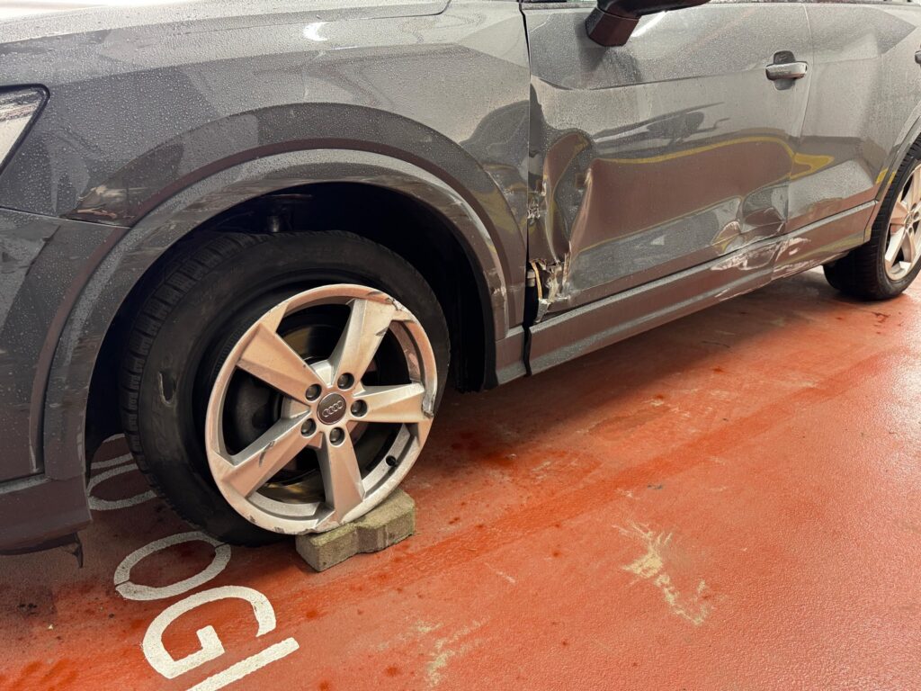 Uszkodzony samochód osobowy z wgniecioną karoserią oraz uszkodzoną felgą stoi na parkingu.