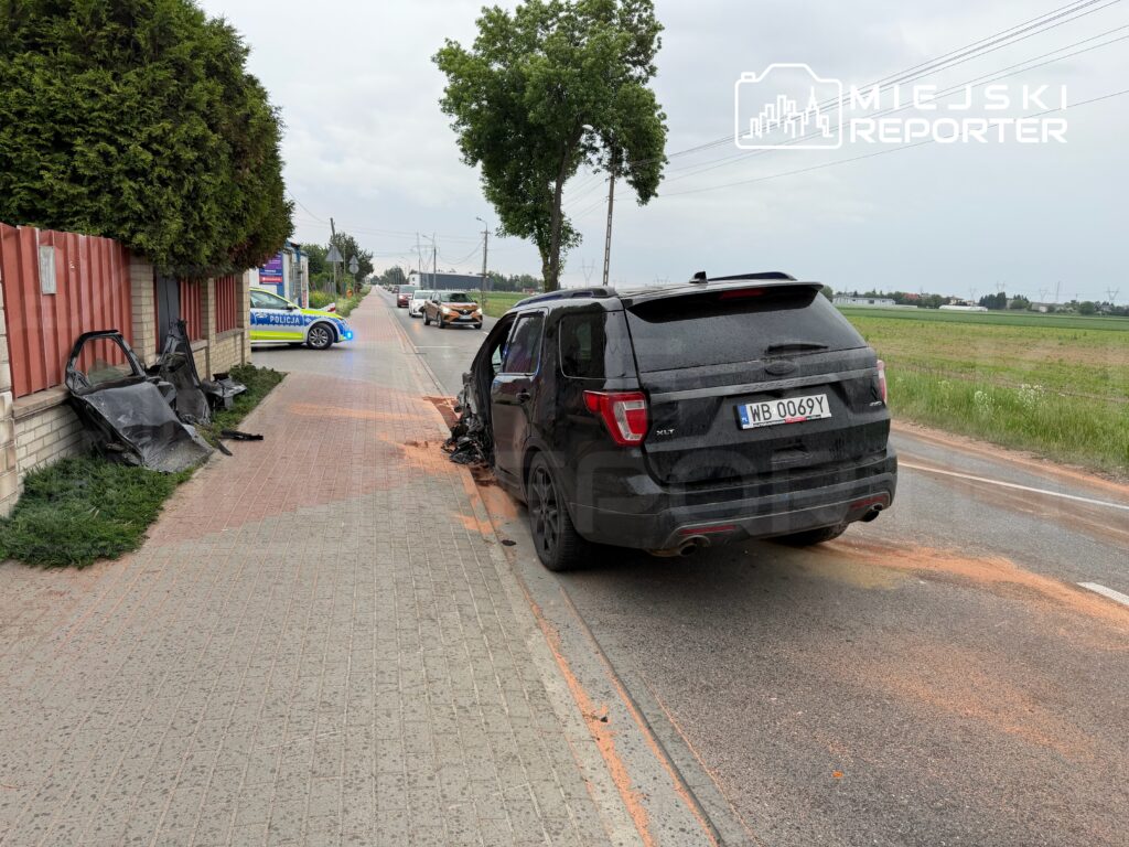 Czarny SUV uszkodzony na poboczu drogi, obok leżące fragmenty wraku oraz radiowóz policyjny z włączonymi sygnałami.