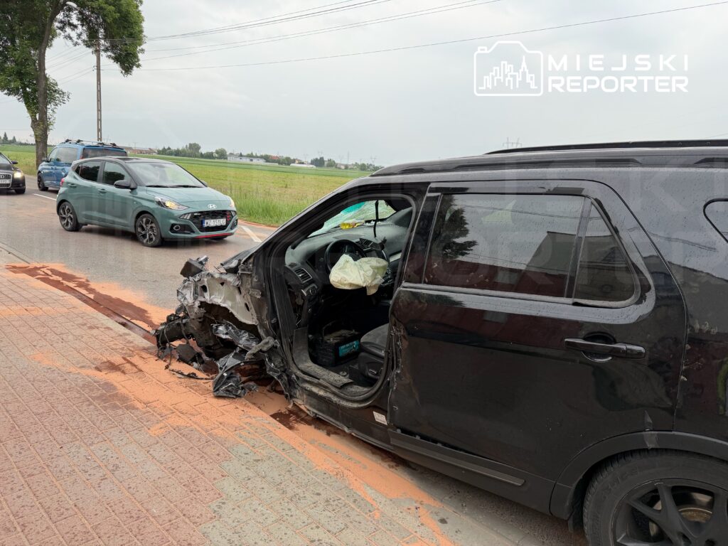 Czarny SUV z uszkodzoną boczną częścią stoi na chodniku, obok widać inne pojazdy na drodze. Na jezdni leży czerwona substancja.