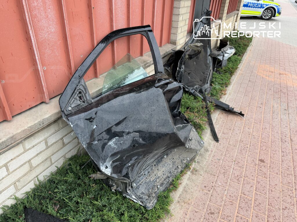 Zniszczona karoseria czarnego samochodu leży na trawie obok budynku, w tle widoczny radiowóz policyjny.