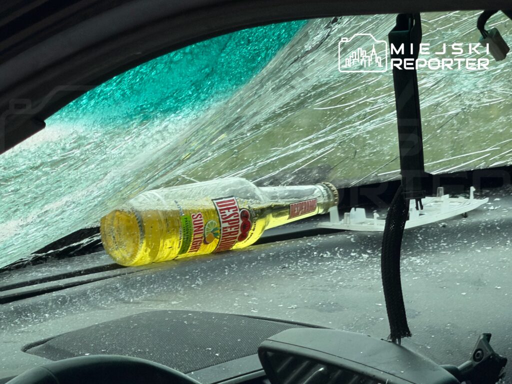 Butelka napoju leży na desce rozdzielczej samochodu z rozbitym przednią szybą, w tle widoczna zniszczona szyba.