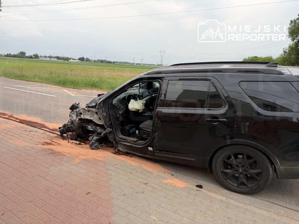 Czarny SUV z uszkodzoną lewą stroną stoi na poboczu drogi, obok widoczne pole i budynki w tle.