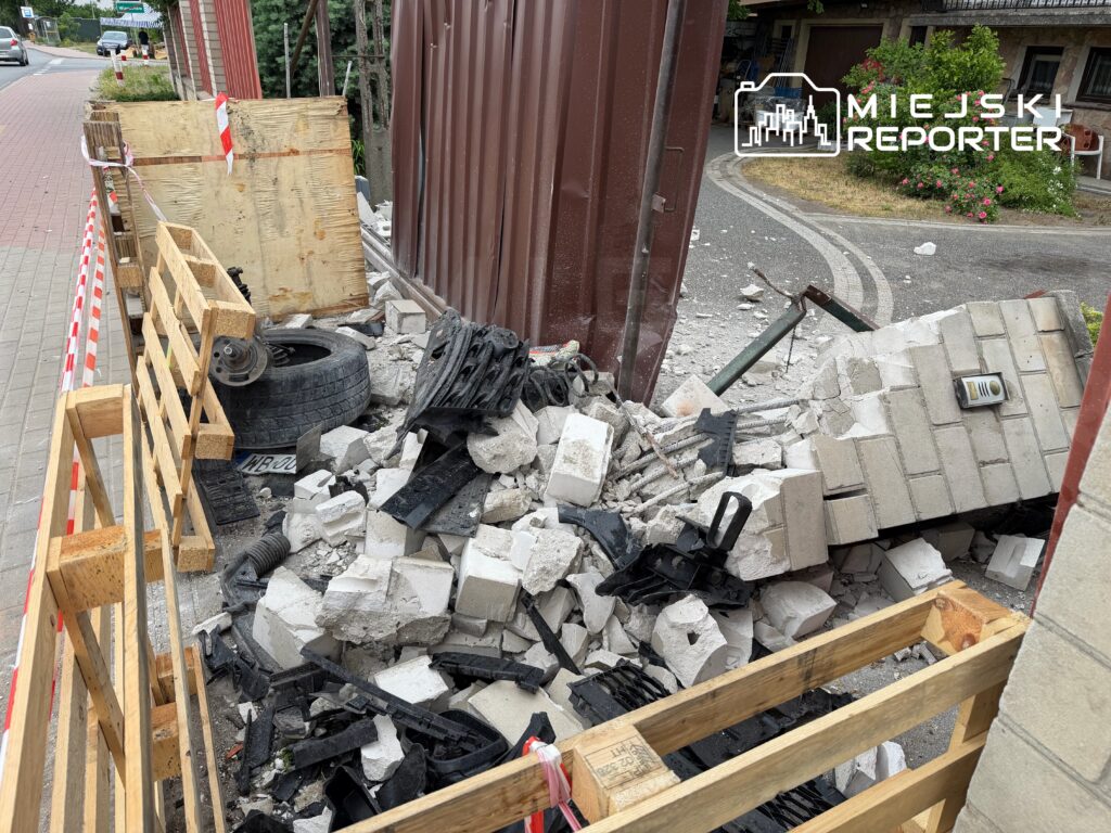 Zniszczona konstrukcja z gruzem, oponą i elementami budowlanymi, otoczona drewnianym ogrodzeniem na chodniku.