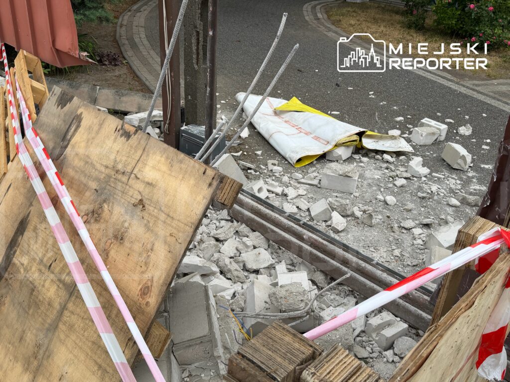 Złom i fragmenty betonu leżą na ziemi obok drewnianych desek i taśmy ostrzegawczej w miejskim otoczeniu.