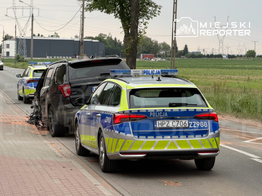 Policyjne samochody zaparkowane przy uszkodzonym pojeździe na poboczu drogi, w tle pola i budynki.