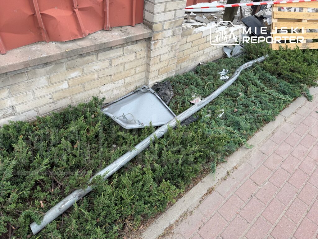 Złamany słupek i uszkodzony element metalowy leżą na zielonym krzewie obok budynku, w tle widoczne cegły i drewno.