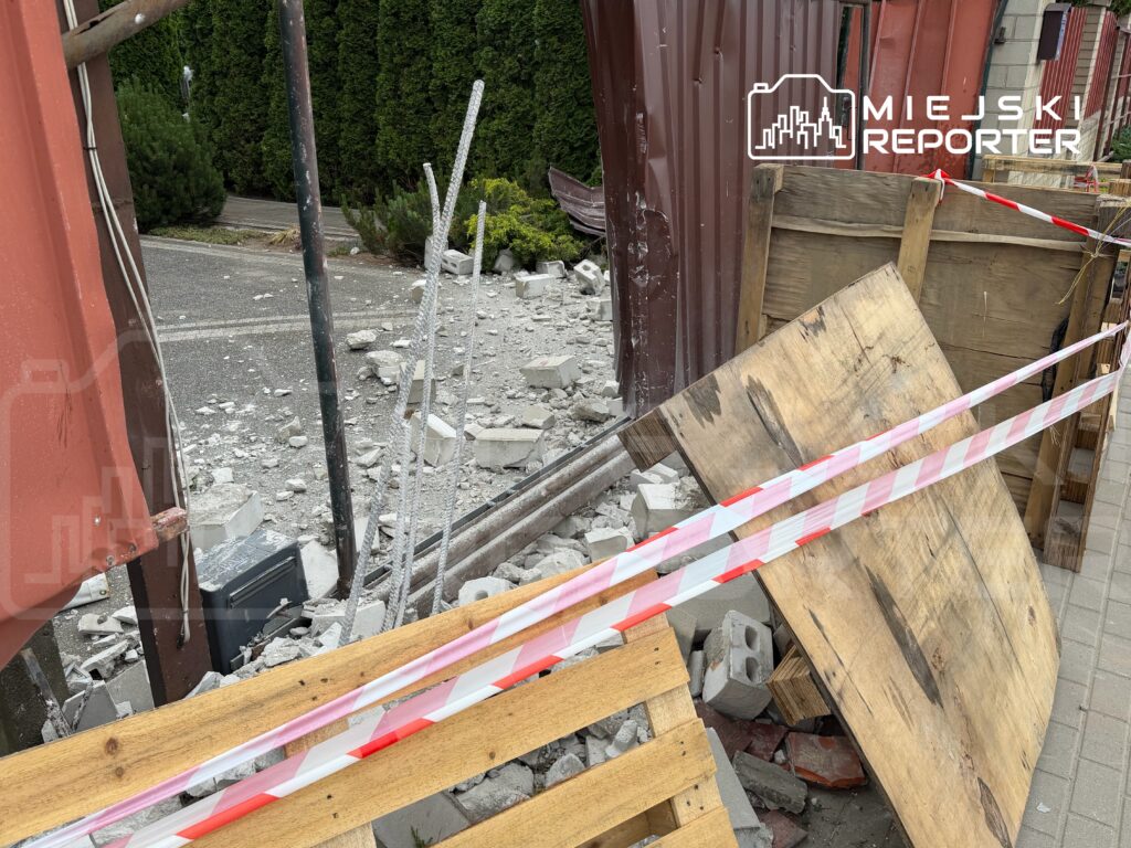 Zniszczony teren budowy z rozrzuconymi fragmentami betonu i drewnianymi paletami, ogrodzony taśmą ostrzegawczą.