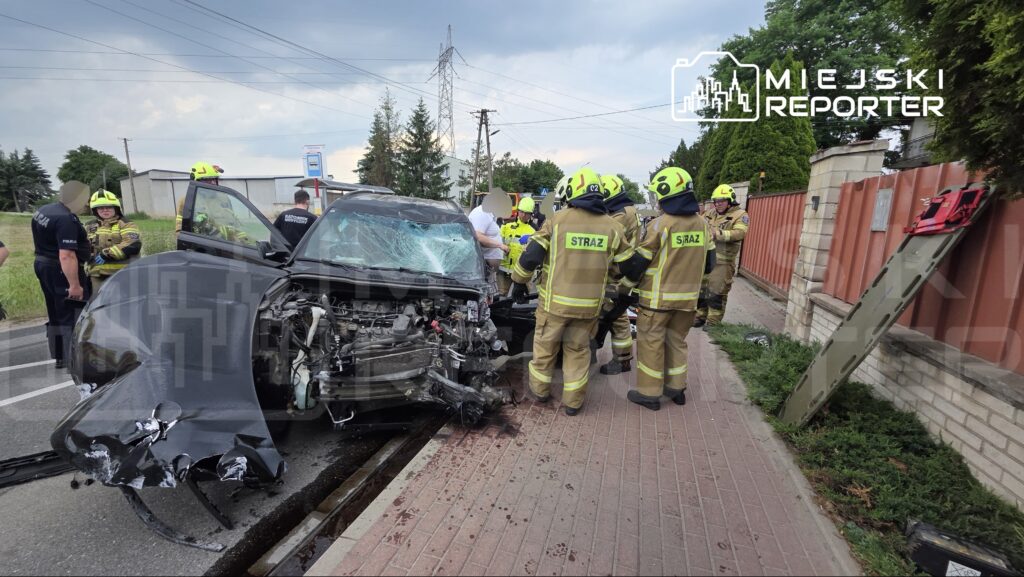 Grupa strażaków w żółtych mundurach udziela pomocy przy rozbitym samochodzie na drodze, obok stoją policjanci.