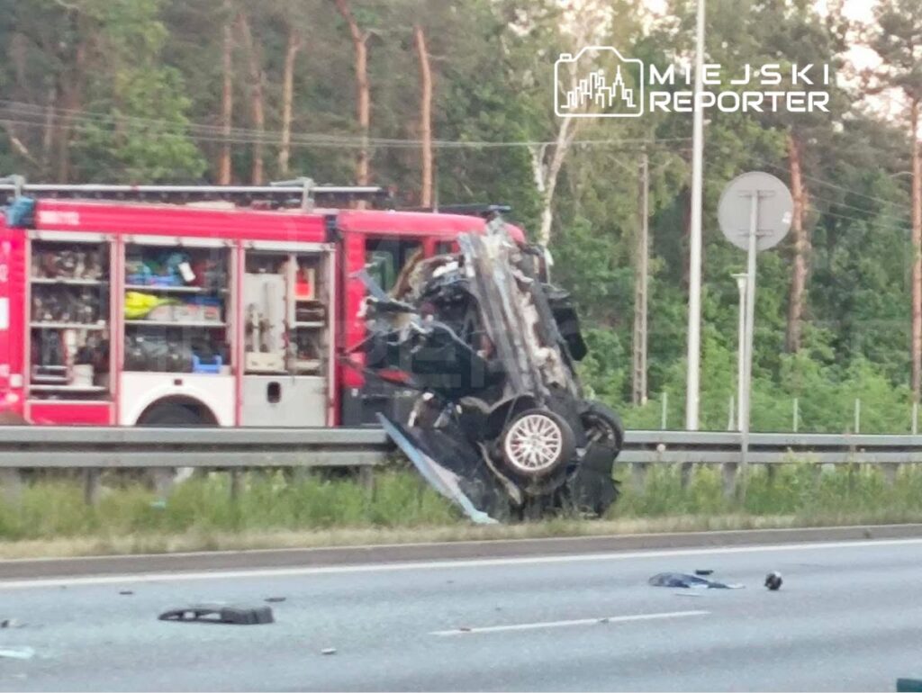 Wypadek drogowy z przewróconym pojazdem na barierze, obok stoi wóz strażacki, na drodze porozrzucane fragmenty samochodu.