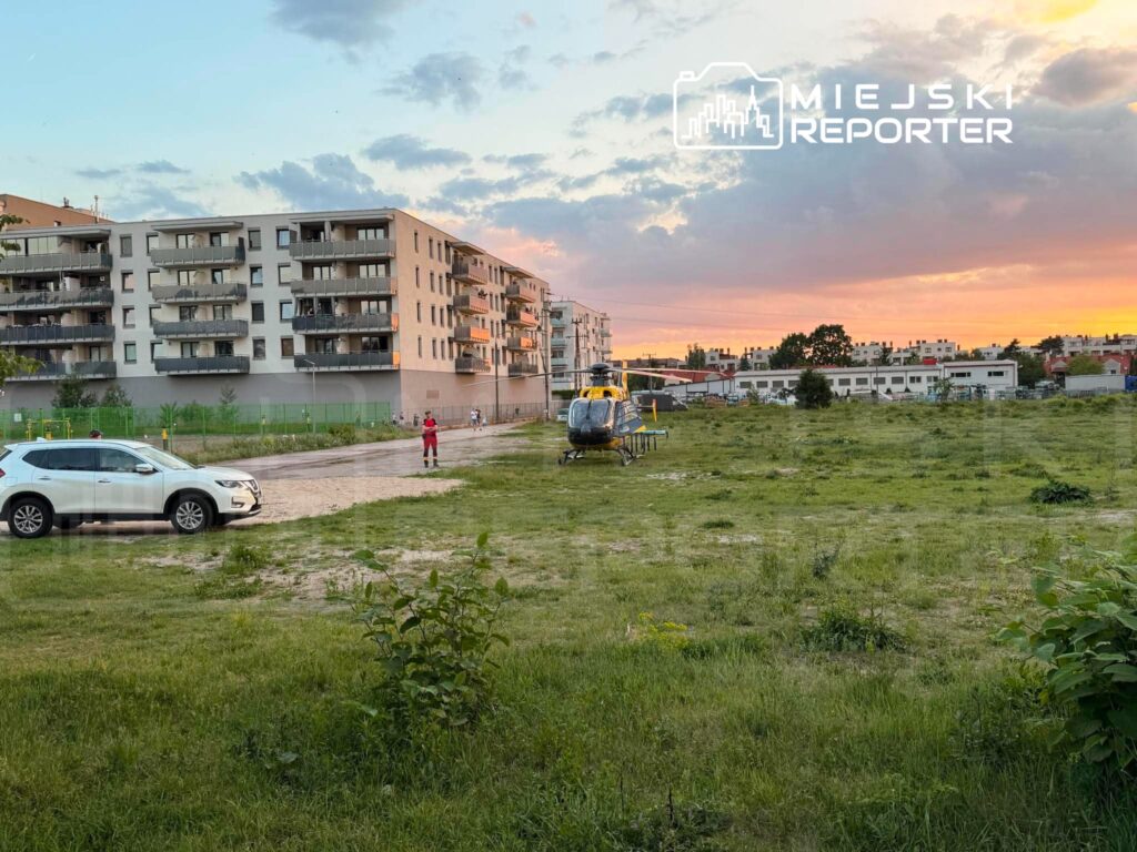 Ratownik w czerwonym stroju stoi obok helikoptera na otwartej przestrzeni, w tle nowoczesne budynki mieszkalne.
