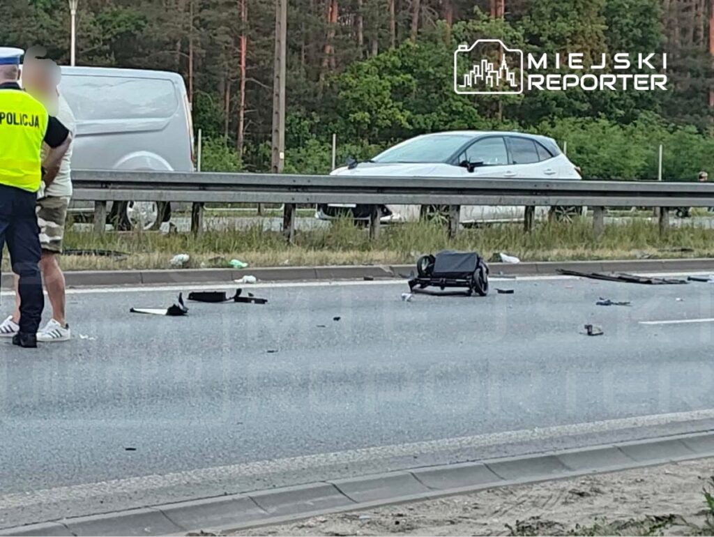 Policjant rozmawia z mężczyzną na drodze, obok leżą fragmenty pojazdu i wózek dziecięcy.